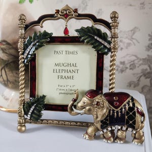 May include: An ornate photo frame with a Mughal elephant design. The frame is gold, burgundy, and black, with jewel accents. The frame holds a 3" x 3" photograph and has the text "Mughal Elephant Frame".