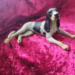 May include: A figurine of a dog, lying down with its legs crossed. The dog has a black and white coat, with long, floppy ears. The figurine is resting on a pink velvet surface.