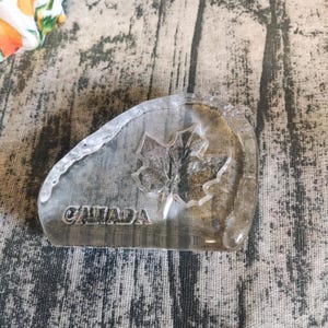 Könnte beinhalten: Ein durchsichtiger Glas-Briefbeschwerer mit einem Ahornblatt-Design und der Aufschrift "CANADA" auf der Unterseite.