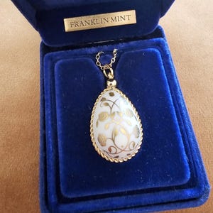 May include: A teardrop-shaped pendant featuring a white surface with gold floral patterns, encased in a twisted gold-tone metal frame. The pendant hangs from a gold-tone chain and is presented inside a blue velvet box labelled FRANKLIN MINT.