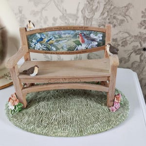 May include: A miniature bench figurine with a painted scene of birds and a landscape. The bench has a wooden appearance with a back panel featuring blue tits and a robin. The base is green, resembling grass, and the bench has floral accents.
