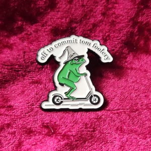 May include: A whimsical enamel pin featuring a green frog wearing a wizard hat and riding a scooter. The pin has the text "off to commit tom foolery" in a white, arched banner above the frog. The pin is outlined in black and set against a pink background.