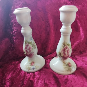 May include: A pair of white ceramic candle holders featuring a floral rose pattern. Each holder has a rounded base, a curved stem, and a cup-shaped top. The roses are pink and red with green leaves and blue accents.