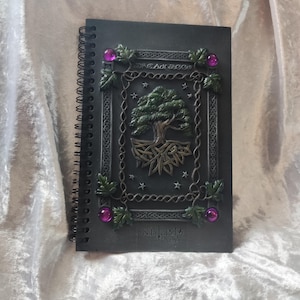 Può includere: Quaderno nero a spirale con copertina in rilievo raffigurante un albero. La copertina include il testo "DREAM BOOK" e "NEHESIS NOW", con dettagli decorativi e gemme viola. Quaderno per scrivere.