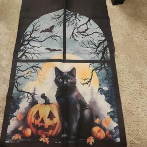 May include: Halloween fabric panel depicting a black cat beside a carved pumpkin. The backdrop shows a full moon, bats, and bare tree branches, creating a spooky scene. The panel is framed by a dark border.