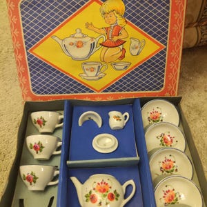 Pode incluir: Um conjunto de chá infantil vintage na sua caixa original. O conjunto inclui uma chaleira, chávenas, pires, um cremeira e um açucareiro, todos decorados com desenhos florais. A caixa apresenta uma ilustração de desenho animado de uma menina.