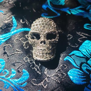 May include: A silver skull ornament, densely covered in clear crystals. The skull features open eye sockets and a detailed jaw. It sits on dark fabric with blue floral embroidery.