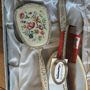 Puede incluir: Un juego de tocador en caja que incluye un espejo de mano, un cepillo para el cabello y un peine. El espejo y el cepillo tienen mangos y respaldos plateados con motivos florales. Una etiqueta en el peine dice HALEX Harmony. El juego está forrado con tela de satén blanco.