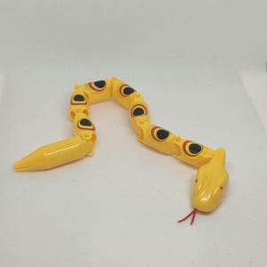 May include: A yellow articulated toy snake with black and red eye details. The snake's body is segmented, allowing for flexible posing. The head features a forked red tongue. The toy is designed to resemble a snake.