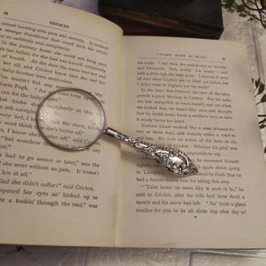 Puede incluir: Un primer plano de una lupa antigua con un mango de plata y un diseño ornamentado. La lupa está apoyada en un libro abierto con texto sobre un bebé y una enfermera. El texto dice "An' then she smiled ever so sweetly, and whispered, 'Bob, darlin', I'm comin'; and with a little sigh she were gone. I knowed it were all over when I called yer to fetch yer father, but I didn't want to frighten yer too much."