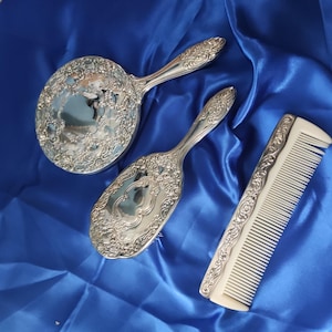 May include: A vintage dressing table set comprising a round hand mirror, a hairbrush, and a comb. The silver-coloured metal items have elaborate floral patterns. The comb has a white handle and teeth. The set is on blue satin fabric.