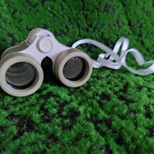 May include: Tan and white compact binoculars with a white neck strap. The binoculars have a textured focus wheel and a central hinge. The lenses are dark, and the body is a light tan color. The binoculars are resting on a bed of green grass.