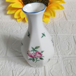 Puede incluir: Un pequeño jarrón de cerámica blanca con un borde azul y un patrón floral. El jarrón tiene un diseño delicado y femenino con rosas rosas y hojas verdes.