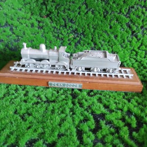 Puede incluir: Un modelo de tren plateado, compuesto por una locomotora y un vagón de carbón, montado sobre una base de madera con vías metálicas. La base tiene una placa con el texto "DEAN GOODS". El tren está sobre un fondo de césped artificial verde.