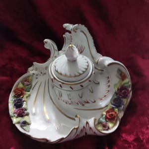 Könnte beinhalten: Ein weißes Porzellan-Tintenfass mit Deckel, verziert mit goldenen Verzierungen und floralen Akzenten. Das Tintenfass ist mit bunten Rosen und aufwendigen Details verziert und schafft ein elegantes und dekoratives Stück.