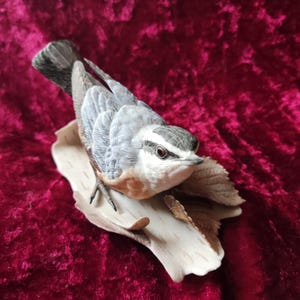 May include: A detailed ceramic bird figurine, featuring a nuthatch perched on a stylized branch. The bird has grey, white, and brown plumage, with a black beak and eye. The branch is a light beige colour. The figurine is set against a deep red background.