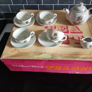 May include: A white children's tea set with a teapot, creamer, four cups, and saucers. Each piece features a gold trim and a small illustration of a child. The set is displayed on a box labeled "Children's China Tea Set."