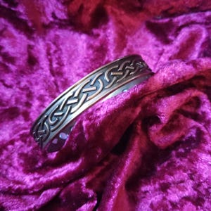 May include: A silver Celtic knot design bracelet with a textured finish. The bracelet is on a burgundy velvet background.