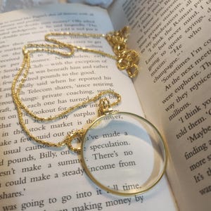 Puede incluir: Un collar con colgante de lupa de color dorado con una lente transparente, apoyado en un libro abierto. La cadena y el marco de la lupa tienen un diseño detallado. Las páginas del libro muestran texto impreso.