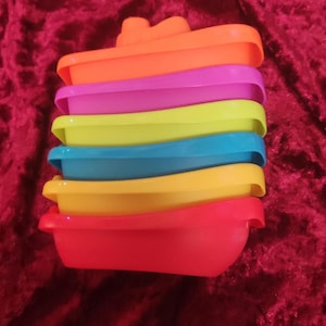 May include: A set of six colourful plastic stacking boats. The boats are red, orange, yellow, blue, green and purple. They are designed to be stacked on top of each other, with the largest boat at the bottom and the smallest boat at the top.