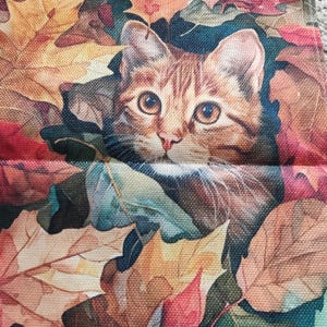 Op de afbeelding: Een close-up van een kattengezicht dat tevoorschijn komt uit een bed van kleurrijke herfstbladeren. De kat heeft bruin en oranje haar met grote, expressieve ogen. De bladeren zijn in tinten rood, oranje, geel en groen.