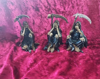 Könnte beinhalten: Drei kleine, sitzende Sensenmann-Figuren. Jede Figur trägt eine schwarze Robe und hält eine Sense. Eine bedeckt ihre Augen, eine ihren Mund und eine ihre Ohren. Die Figuren stehen auf rotem Samt.
