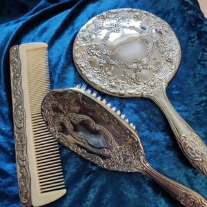 May include: A vintage vanity set featuring a round hand mirror, a hairbrush, and a comb. The set has ornate silver-colored detailing with floral patterns. The comb and brush have ivory-colored handles and bristles, set against a deep blue velvet background.