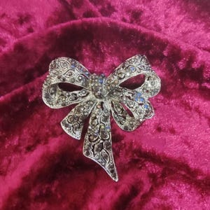 Puede incluir: Un broche de lazo plateado con un diseño detallado y diamantes de imitación iridiscentes.