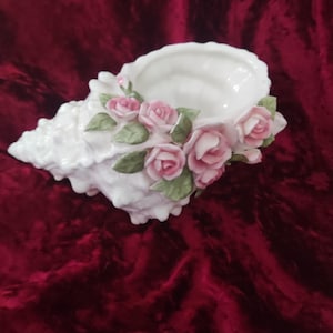 May include: A white ceramic seashell-shaped decorative bowl. The bowl is decorated with pink roses and green leaves. The seashell has a textured surface and a smooth, white interior. The bowl is a decorative item.