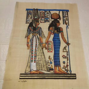 May include: A hand-painted papyrus scroll depicting two Egyptian goddesses standing side-by-side. The goddess on the left wears a white and gold dress with a red and green sash. The goddess on the right wears a blue dress with a gold belt. Both goddesses have elaborate headdresses and jewellery.