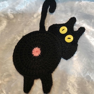 May include: Black crocheted cat-shaped hot pad with yellow button eyes and a pink crocheted tail.