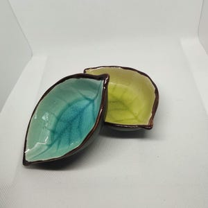 Puede incluir: Dos pequeños cuencos de cerámica con forma de hojas. Un cuenco es azul verdoso con un borde marrón y el otro es amarillo con un borde marrón.