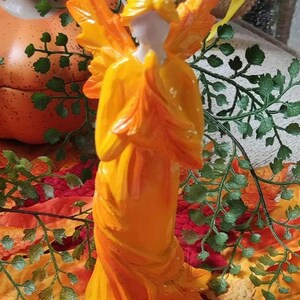 May include: A vibrant, orange-yellow angel figurine with outstretched wings and clasped hands. The statue has a white face and is set against a backdrop of fall foliage and a pumpkin, evoking a seasonal or harvest theme.
