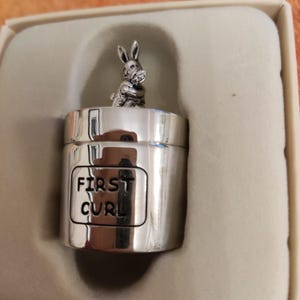 Könnte beinhalten: Eine silberfarbene Andenkenbox mit einer Hasenfigur auf der Oberseite. Auf der Box steht "FIRST CURL". Die Box wird in einer Geschenkbox in neutralen Farbtönen präsentiert.