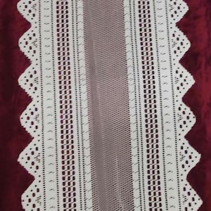 Puede incluir: Un camino de mesa de crochet blanco con un patrón geométrico. El camino de mesa tiene un borde festoneado y está colocado sobre una superficie burdeos.