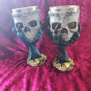 Könnte beinhalten: Zwei dekorative Becher mit Totenkopf-Design. Die Becher haben einen silberfarbenen Rand und ein detailliertes Totenkopfgesicht. Der Stiel ist ein Drachenklauen-Design mit goldfarbener Basis. Die Becher stehen auf rotem Samt.