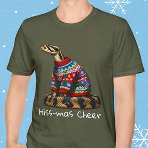 May include: Olive green t-shirt featuring a snake wearing a colorful Christmas sweater. The snake is coiled and the text "Hiss-mas Cheer" is printed below. The background is a light blue with white snowflakes.