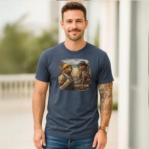 Może przedstawiać: Krótki rękaw t-shirt w kolorze melanżowego granatu z grafiką przedstawiającą dwa bobry ubrane jak robotnicy budowlani przy tamie Hoovera. Jeden bóbr ma żółty kask i ogrodniczki, drugi fedorę i garnitur. Napis na grafice brzmi Hoover Dam 1936.
