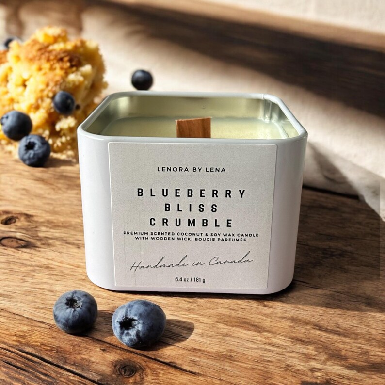 K&ouml;nnte beinhalten: Ein wei&szlig;es Kerzengef&auml;&szlig; mit einem Holzdeckel. Das Etikett lautet "LENORA BY LENA BLUEBERRY BLISS CRUMBLE PREMIUM SCENTED COCONUT & SOY WAX CANDLE WITH WOODEN WICKI BOUGIE PARFUMEE Handmade in Canada 8.4 oz/181 g".