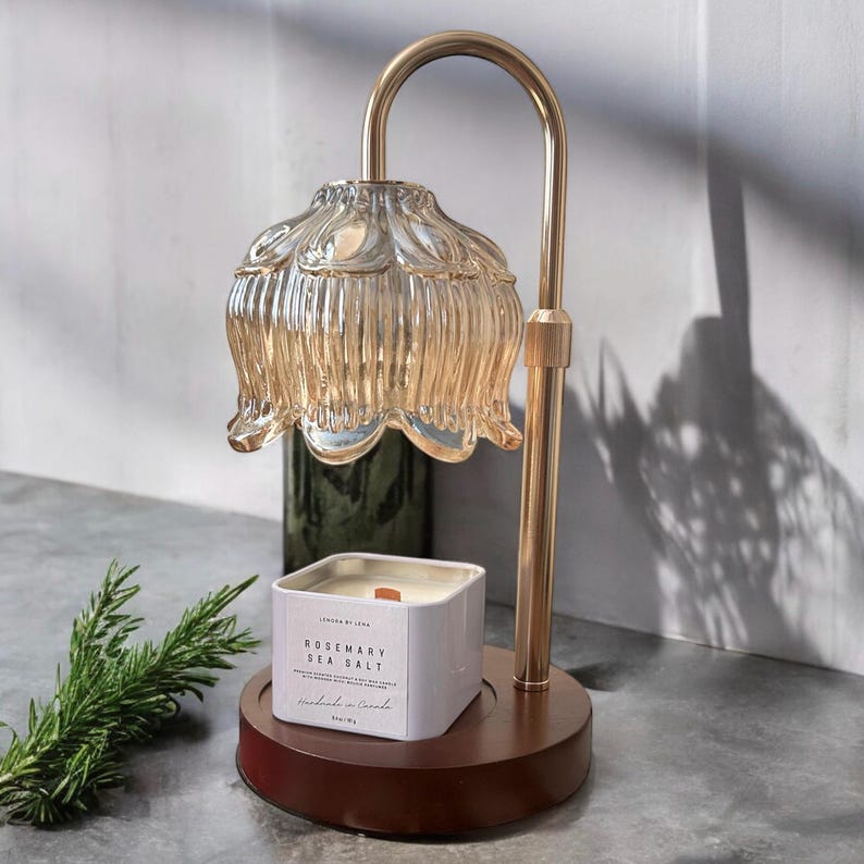 K&ouml;nnte beinhalten: Ein goldfarbener Kerzenw&auml;rmer aus Metall mit einem glasernen, blumenf&ouml;rmigen Schirm. Eine wei&szlig;e quadratische Kerze mit dem Text "Rosemary Sea Salt" befindet sich auf der Basis des W&auml;rmers.