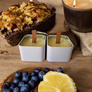 K&ouml;nnte beinhalten: Zwei wei&szlig;e quadratische Kerzen mit Holzdochten, eine Zitronenscheibe, Blaubeeren und ein gebackener Nachtisch mit Streuseln.