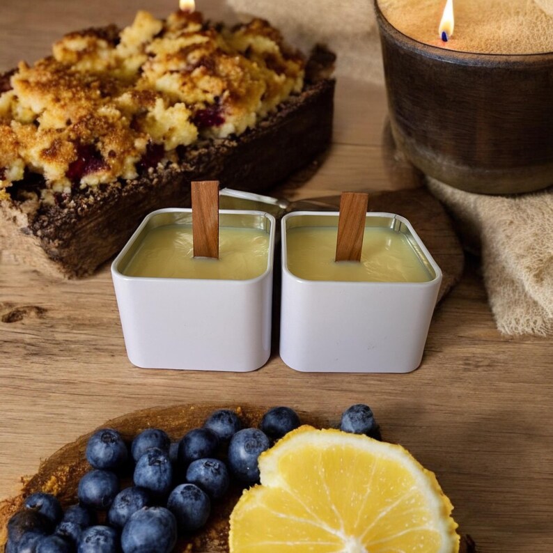 K&ouml;nnte beinhalten: Zwei wei&szlig;e quadratische Kerzen mit Holzdochten, eine Zitronenscheibe, Blaubeeren und ein gebackener Nachtisch mit Streuseln.