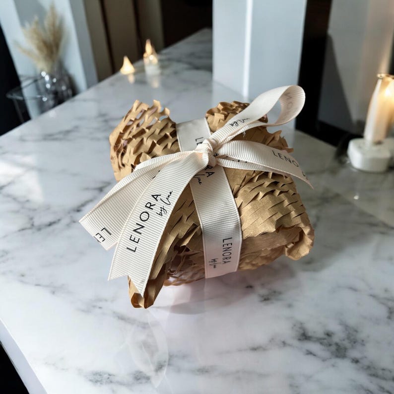 K&ouml;nnte beinhalten: Eine kleine Geschenkbox, die in braunes Papier eingewickelt ist und ein wei&szlig;es Band mit Schleife hat. Das Band tr&auml;gt den Aufdruck "LENORA by Uma".