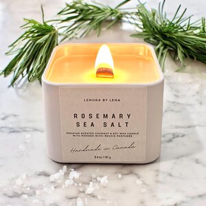K&ouml;nnte beinhalten: Eine wei&szlig;e quadratische Kerze mit einem Holzdocht, beschriftet mit "Rosemary Sea Salt" und "Lenora by Lena". Die Kerze besteht aus Kokosnuss- und Sojawachs und wird in Kanada handgefertigt. Sie wiegt 8,4 Unzen oder 181 Gramm.