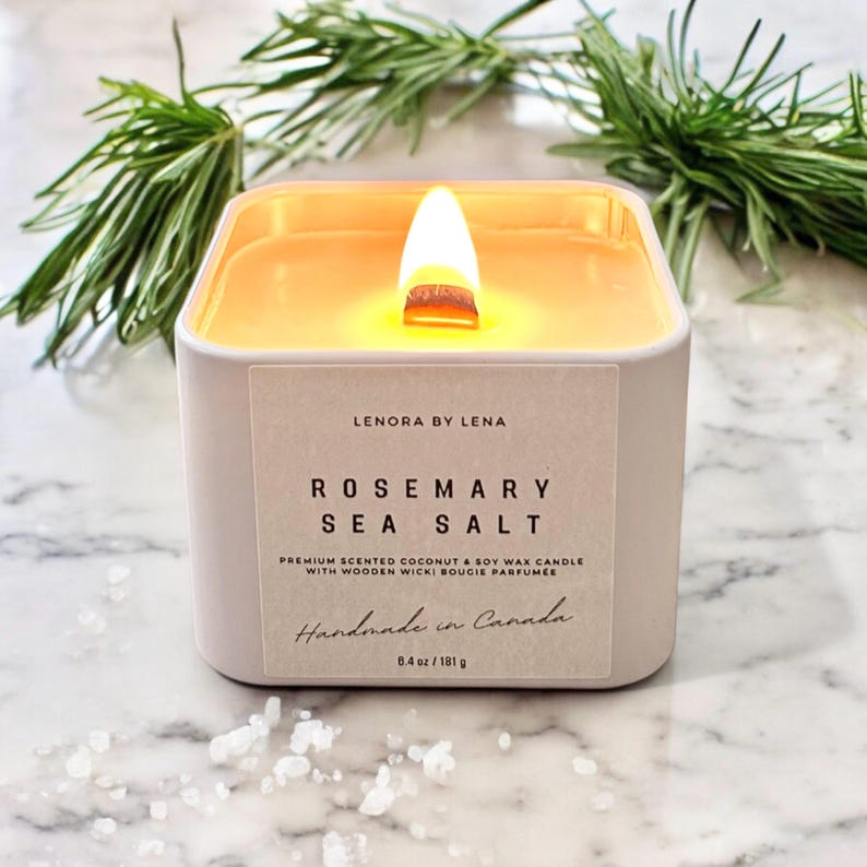 K&ouml;nnte beinhalten: Eine wei&szlig;e quadratische Kerze mit einem Holzdocht, beschriftet mit "Rosemary Sea Salt" und "Lenora by Lena". Die Kerze besteht aus Kokosnuss- und Sojawachs und wird in Kanada handgefertigt. Sie wiegt 8,4 Unzen oder 181 Gramm.