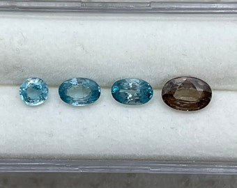 May include: Four oval-shaped gemstones in a white display case. The gemstones are a light blue, a darker blue, a light blue, and a brown.