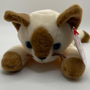 Puede incluir: Un peluche de gato siamés con pelaje color crema y marcas marrones en las orejas, patas y cara. El gato tiene ojos azules y una etiqueta roja. El juguete está tumbado.