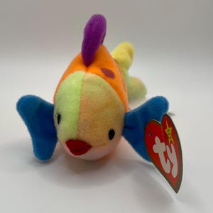 Puede incluir: Un juguete de peluche con forma de pez colorido con detalles en naranja, amarillo, verde y azul. El pez tiene una aleta morada y una boca roja. Una etiqueta roja en forma de corazón con el texto "ty" está adjunta.