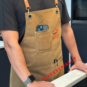 May include: A brown canvas apron with leather straps and details. The apron features a front pocket containing a phone, a pen, and two leather patches. The apron is worn by a person wearing a black top.
