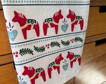 May include: A white kitchen towel featuring a repeating pattern of red Dala horses, light blue hearts, and green pine branches with red berries. The design is organized in horizontal bands separated by rows of small light blue dots.